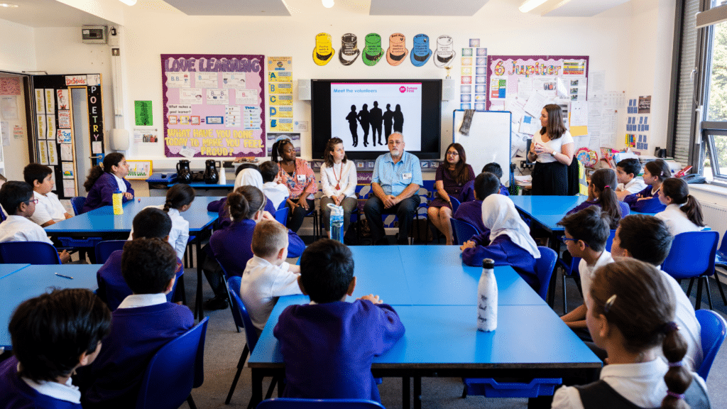 a group of school children sat at classroom tables look up to four adults sat at the front of the classroom